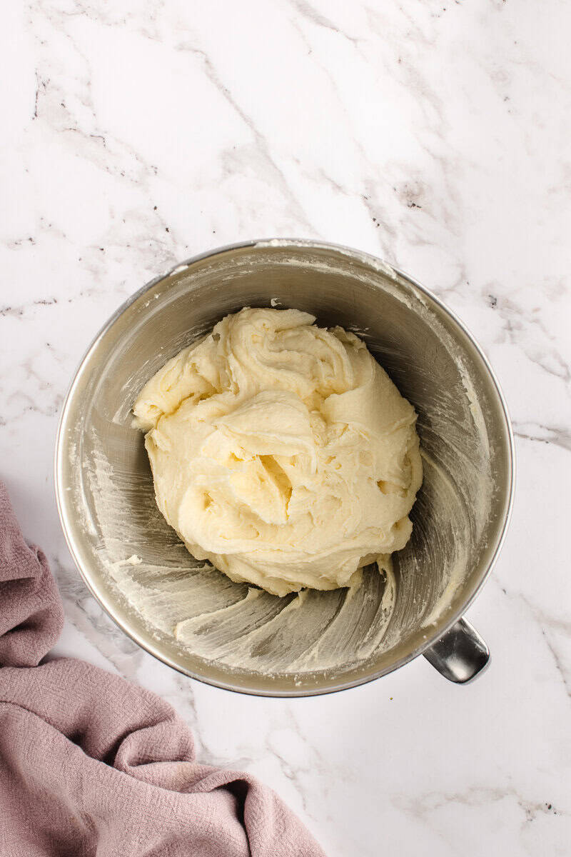 A stainless steel bowl containing creamy, mixed dough on a marble counter with a pink cloth nearby.